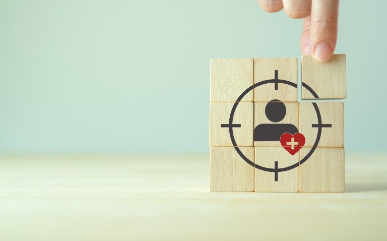 A stack of nine wooden blocks in a square with a hand putting the top right block into place and a silhouette of a person in the middle with a red heart shape overlapped with a white medical cross in the centre and a target shape around them to illustrate a new frontier in precision medicine for complex disease