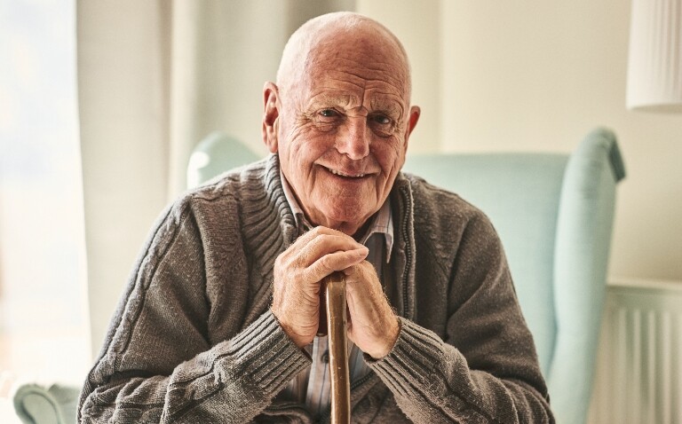 An old man sitting on a chair at home with his hands clasped over a walking stick to represent a risk of falls as part of a new AI tool aiming to reduce hospital admissions