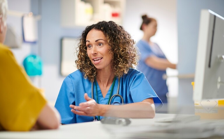 A female doctor in scrubs sat having a consultation with a patient in a yellow tshirt and a computer on the desk next to them and a colleague in the background to illustrate the launch of Microsoft's ambient voice technology tool in the UK