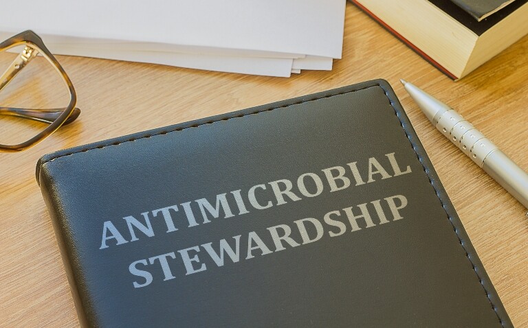 A black leather folder with the words antimicrobial stewardship on the front sat on a desk with a notepad