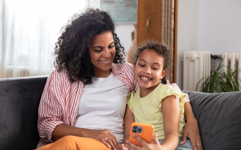 A mother and daughter sat on a sofa smiling while looking at a smartphone being held by the daughter to illustrate the discovery that an at-home monitoring app can improve asthma control and reduce hospitalisations in children
