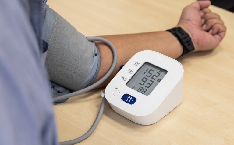 A close up of a man wearing a blue top with his arm in a blood pressure cuff resting on a table with the digital display of the blood pressure monitor showing a reading of 152/93 to illustrate the finding that baxdrostat significantly reduces hard-to-control hypertension