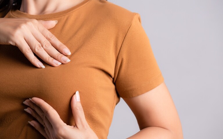 A close up of a woman in an orange short-sleeved t-shirt checking her breasts for signs and symptoms of breast cancer to illustrate the need to encourage regular breast checking from two Breast Cancer Awareness Month surveys