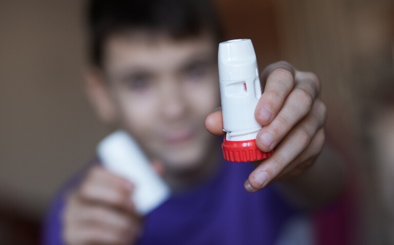 A young boy wearing a purple tshirt and reaching his arms forwards holding a white and red Turbohaler inhaler with the main part in one hand and the cover in the other hand to illustrate the finding that a combined budesonide-formoterol inhaler cuts asthma attacks in children
