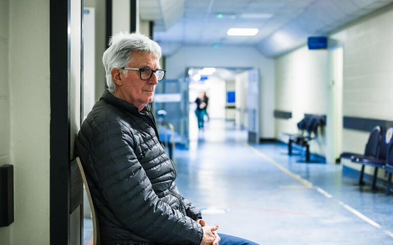 An older man sitting on a chair in a hospital corridor waiting for his appointment to illustrate calls for updated prostate cancer guidance after overtreatment identified