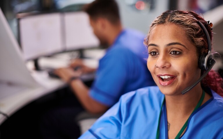 A healthcare professional wearing blue scrubs sat at a computer and talking on a headset with a colleague in the background looking at multiple computer screens to illustrate clinical areas being prioritised for technology expansion following the NHS 10-year plan launch