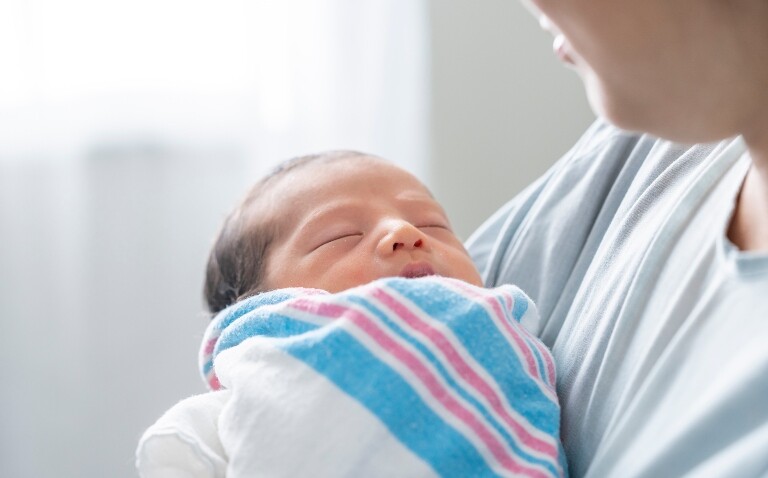 A close-up of a baby wrapped in a blanket being held by its mother to illustrate research that has identified a higher risk of infection in children born to women with obesity