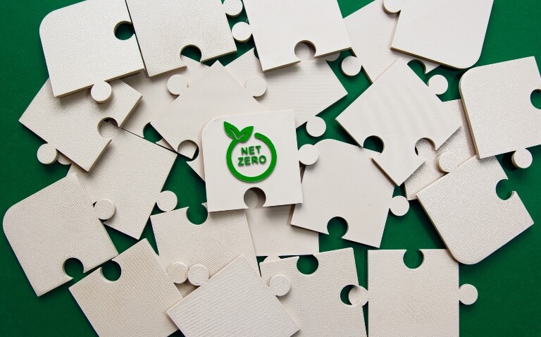 A pile of off-white jigsaw pieces on a green background with the words net zero in a circle with two leaves on the edge on the top piece to illustrate UCLH's sustainability progress and advice for clinicians to embrace their Trust green plan