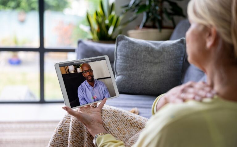 A woman having a video call with her doctor while sat on a sofa at home and holding her shoulder