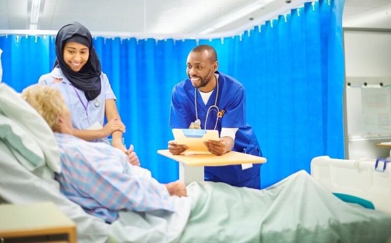 A smiling nurse and a doctor talking to an older woman at her hospital bedside with blue curtains drawn to illustrate the largest ever NHS budget announcement as part of the Government's spending review