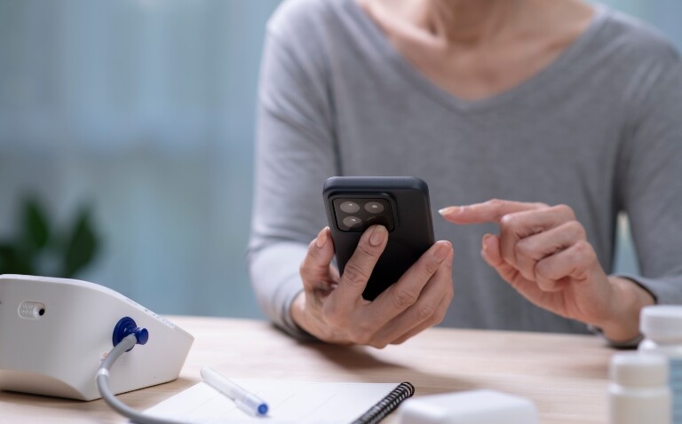 A older woman sitting at a table with a blood pressure monitor and a pen and notebook holding a smartphone to illustrate the success and expansion of Leicester's AF virtual ward project