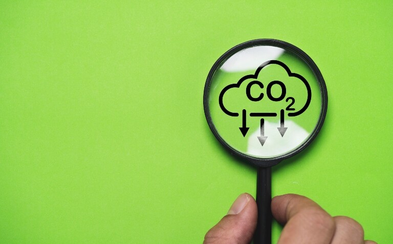 Hand holding a magnifying glass over a reducing CO2 reducing icon on a green background to illustrate a low-carbon COPD inhaler receiving MHRA approval