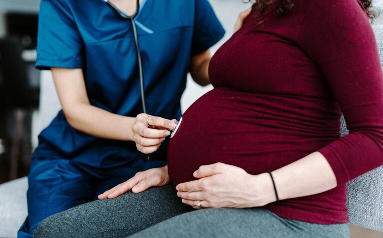 A doctor in scrubs using a stethoscope to listen to a woman's pregnant abdomen to illustrate that maternal anaemia has been linked with congenital heart disease in babies