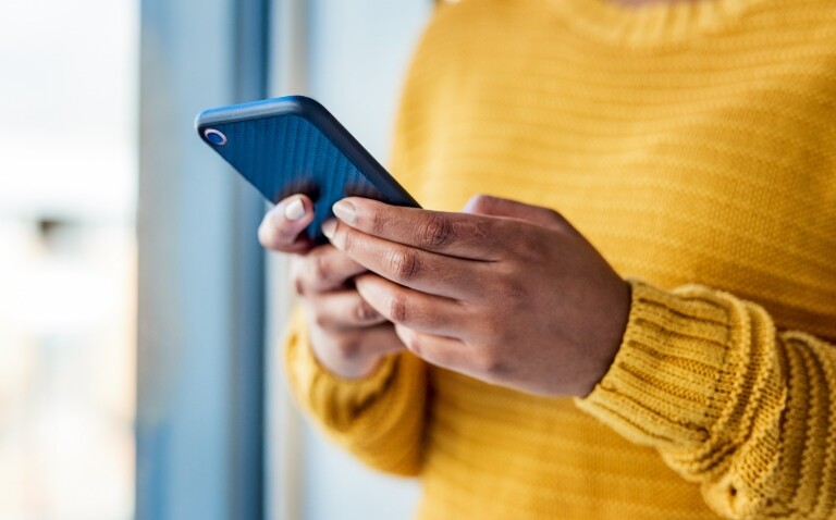 Close up of a woman in a yellow jumper looking at a smartphone to illustrate use of the NHS App saving clinical hours and being adopted by the majority of UK hospitals