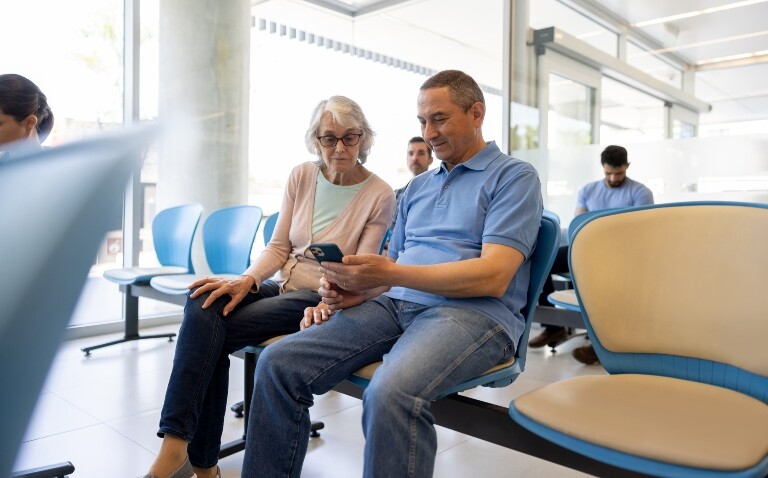 Older woman with her son looking at a smartphone in a hospital waiting room to illustrate the announcement that the NHS App will become the go-to patient communication method within three years