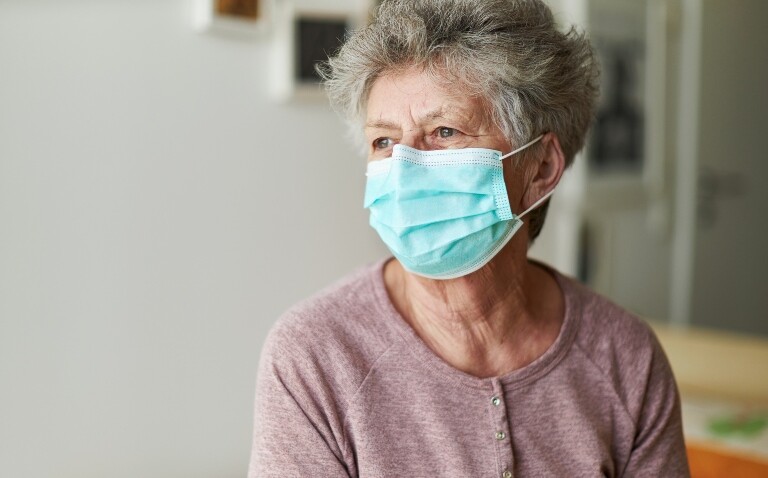 A head and shoulders shot of an older woman wearing a surgical mask and a pink cardigan to illustrate NICE removing eligibility for nirmatrelvir plus ritonavir for some at-risk groups
