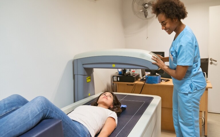 Smiling healthcare worker is looking at young woman lying on a bone density scanner to illustrate the DHSC announcing the location of 13 new scanners