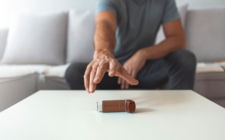Male hand reaches for a brown asthma inhaler while sitting on a sofa to represent optimum timing for beclomethasone inhaler use