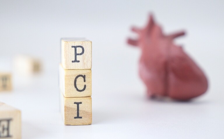 Three wooden blocks stacked on top of each other with the letters PCI on each one and a model of a human heart blurred in the background to illustrate the finding that early aspirin discontinuation and P2Y12 inhibitor monotherapy has benefits in low-risk MI patients after PCI