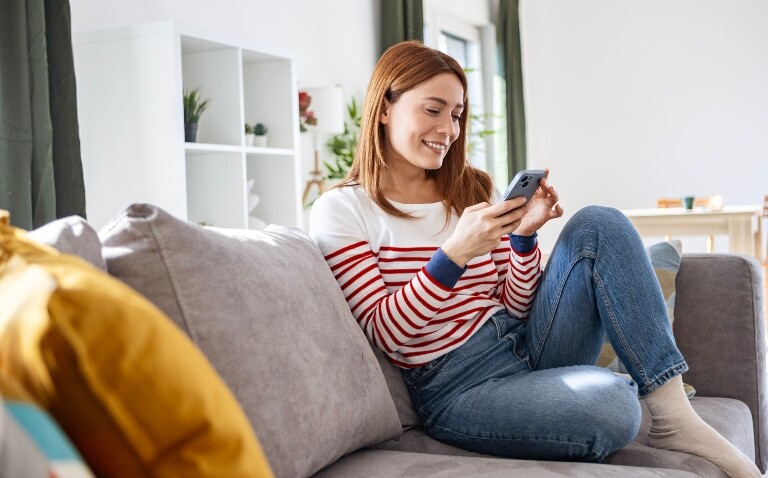 A young woman sat on a sofa looking at a smartphone and smiling to illustrate the announcement of patient access to trusted health information via the NHS App being a focus of the NHS 10-year plan