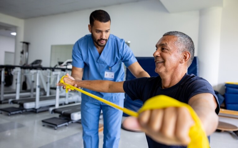 Older man doing physical therapy exercises using a stretch band with the assistance of a healthcare professional to illustrate personalised rehabilitation for multimorbidity such as hypertension and asthma showing early promise