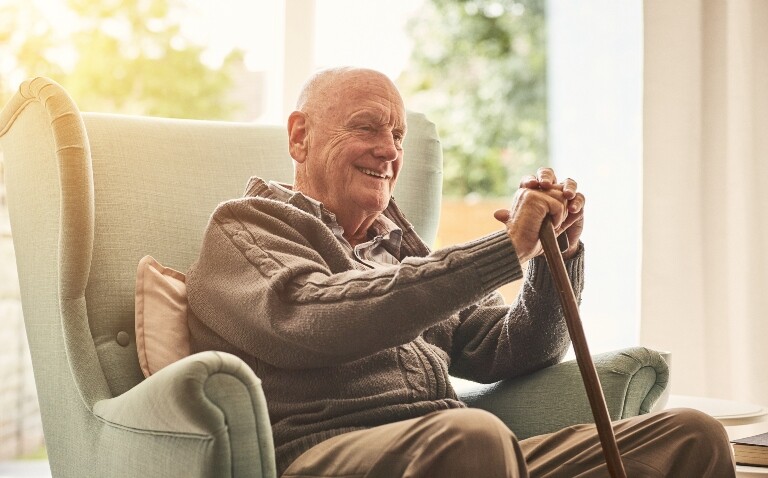 An elderly man sitting in a wingback chair holding a walking stick and smiling