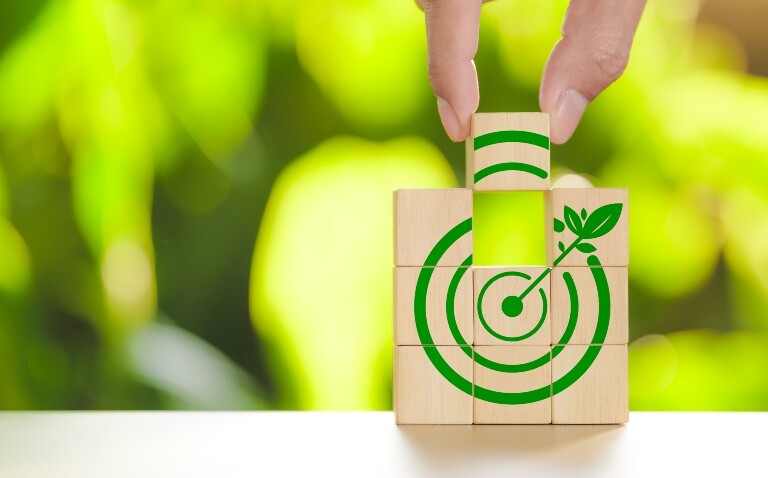 Eight wooden blocks stacked with a green target and dart shape on the front with two fingers placing the ninth block into the centre of the stack and blurred green leaves behind to illustrate positive progress being made towards NHS interim carbon footprint targets