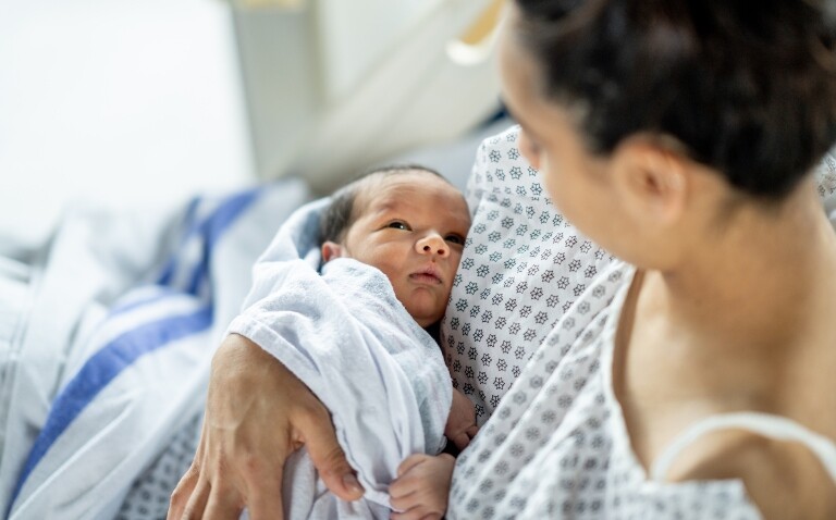 A close up of a newborn baby wrapped in a blanket being held by its mother who is wearing a hospital gown and sitting up in bed to illustrate research from Professor Nigel Field into the gut microbiome and linking it with the prevention of childhood viral infections