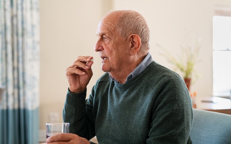 An old man sitting on a sofa at home taking a tablet and holding a glass of water to illustrate the real-world impact of medication adherence in pulmonary hypertension