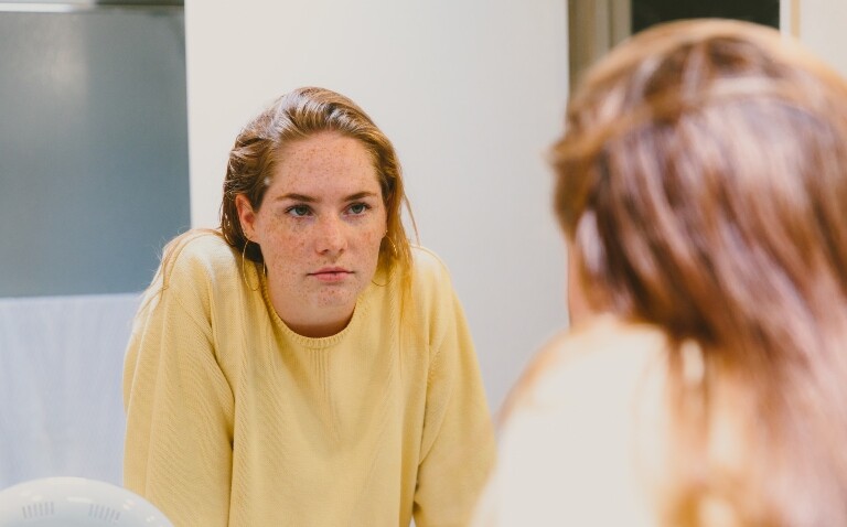 A teenage girl with ginger hair and freckles wearing a yellow jumper looking into a mirror to illustrate the iCARE Health Hub mirror supporting AYA cancer survivors