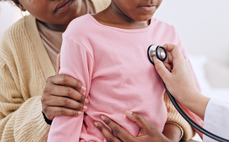 A young girl sitting on her mum's lap while a doctor listens to her chest with a stethoscope to illustrate the risk of cardiac complications in children after Covid infection and vaccination