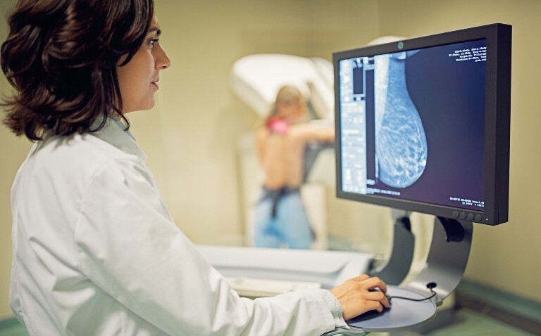 A woman in the background having a mammogram with a healthcare professional standing at a screen in the foreground looking at the results to illustrate the finding that risk of breast cancer death is 40% higher if a woman's first mammogram is missed