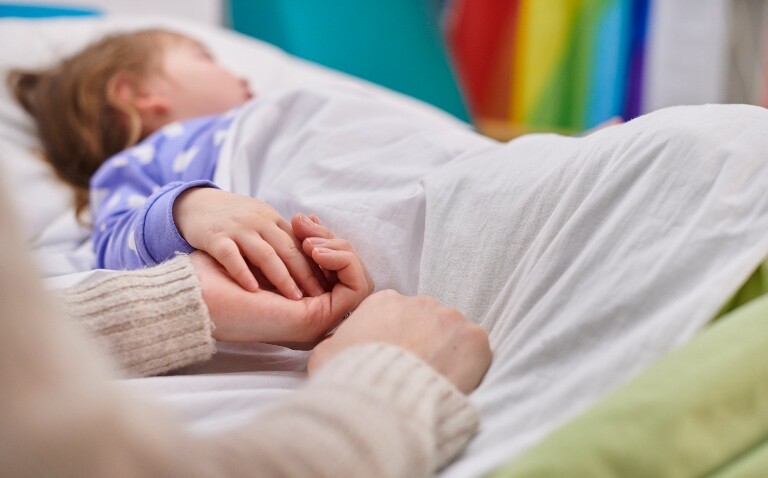 A child laying in a hospital bed with their mum holding their hand to represent strain on the paediatric workforce posing serious risk to child health as the RCPCH calls for action