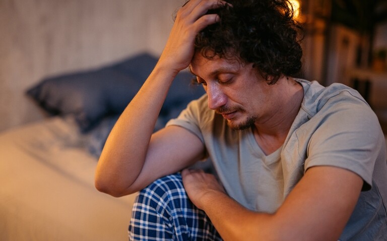 A man sitting on a bed at night with his head in his hand to illustrate sleep disorders potentially being a predictive marker of future neurodegeneration