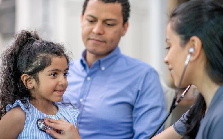 A little girl sitting on her dad's knee while a doctor listens to her lungs through a stethoscope to illustrate the need to take action on child asthma and anaphylaxis deaths