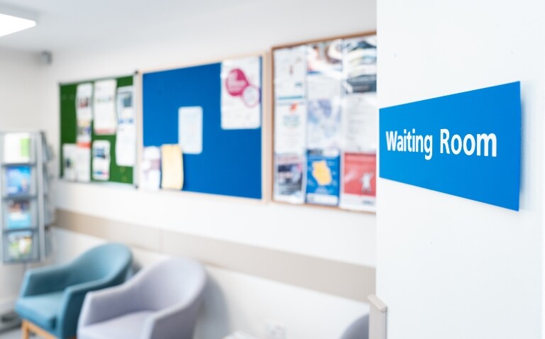 A view into a hospital waiting room with empty chairs and noticeboards on the walls and a blue waiting room sign on the door to illustrate trust leaders being forced to scale back services and clinical staff due to financial pressures
