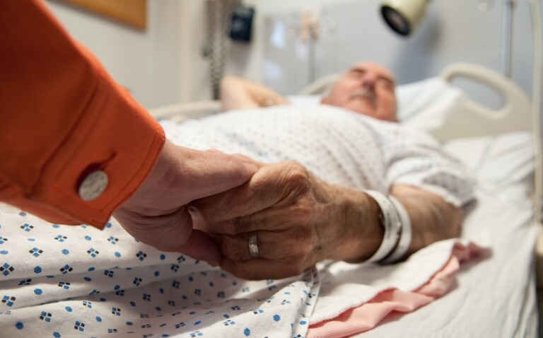 An older man lies in a hospital bed holding hands with a visitor