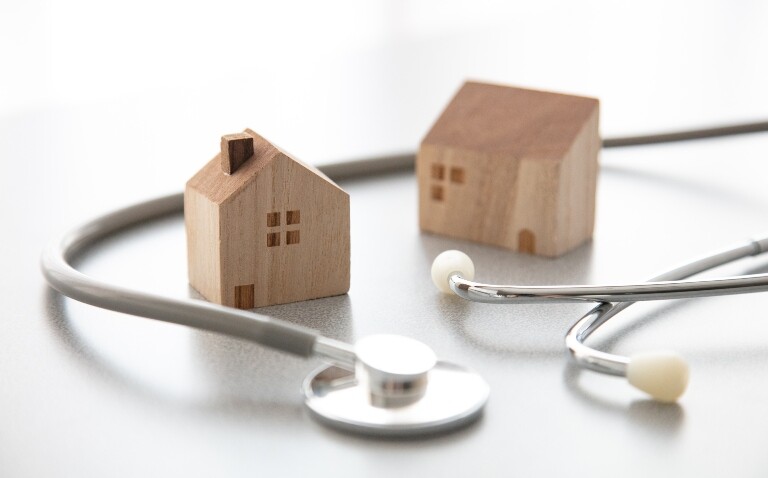 A wooden house and a separate wooden building with a stethoscope circled around them to represent the new NHS 10-year plan moving care from hospitals to the community and closer to patients' homes