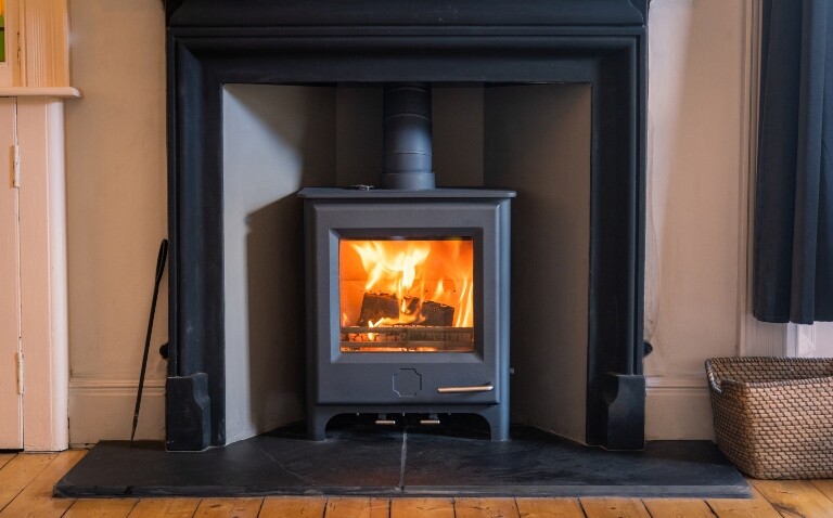 A black fireplace with a grey wood-burning stove in the centre with a fire visible through a glass screen and a log basket on the left-hand side and fire tools on the left to illustrate new research showing that log burners expedite lung function decline