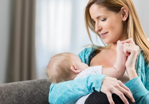 A woman wearing a blue cardigan sitting on a grey sofa and breastfeeding her baby to illustrate the finding that human milk-gut microbiome interactions are linked to childhood blood pressure
