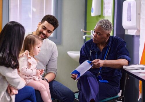 A female doctor sitting with two parents and their daughter in a hospital and showing them a leaflet to illustrate the acceleration of childhood cancer diagnosis through the Child Cancer Smart awareness campaign