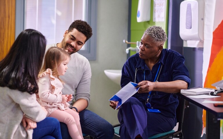A female doctor sitting with two parents and their daughter in a hospital and showing them a leaflet to illustrate the acceleration of childhood cancer diagnosis through the Child Cancer Smart awareness campaign