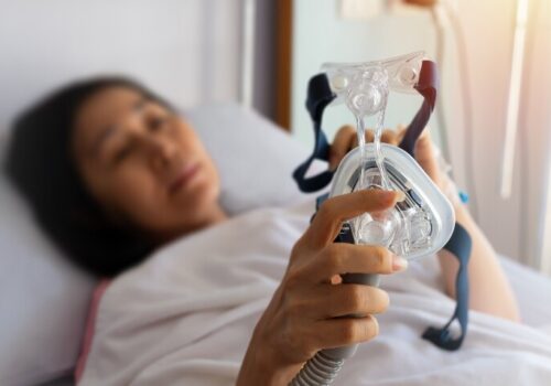A woman lying in a bed holding a CPAP mask to illustrate the finding that action on OSA diagnosis and treatment is required as economic and clinical burden revealed