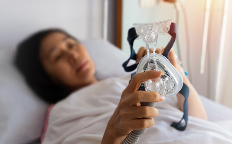 A woman lying in a bed holding a CPAP mask to illustrate the finding that action on OSA diagnosis and treatment is required as economic and clinical burden revealed