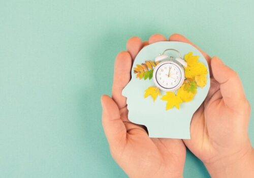 two hands on a light green background holding a human head shape piece of card with a clock showing 2 o'clock surrounded by autumn leaves to illustrate the autumn daylight saving clock change being linked to reduced mental and physical health issues and reduced demand on the health system