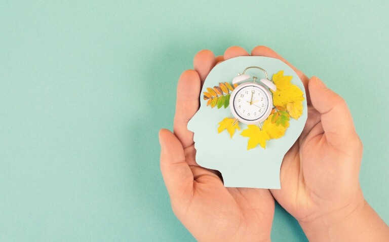 two hands on a light green background holding a human head shape piece of card with a clock showing 2 o'clock surrounded by autumn leaves to illustrate the autumn daylight saving clock change being linked to reduced mental and physical health issues and reduced demand on the health system