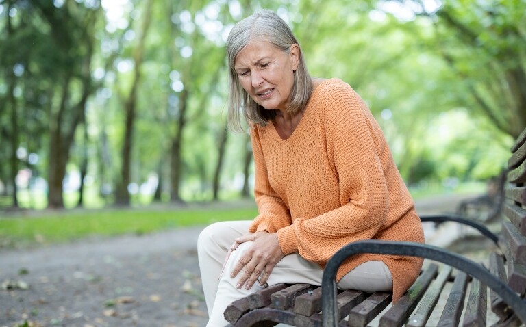 Senior woman feeling knee pain sitting on bench in park