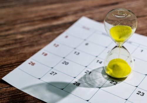 A glass hourglass with yellow sand sitting on a page of a calendar on a wooden table to illustrate the benefits of early biologic initiation in psoriasis