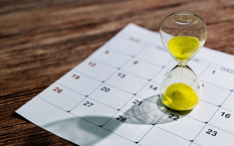 A glass hourglass with yellow sand sitting on a page of a calendar on a wooden table to illustrate the benefits of early biologic initiation in psoriasis