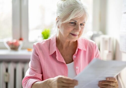 A woman wearing a pink shirt and white tshirt sat at a kitchen table and reading a letter to illustrate a mammogram invitation